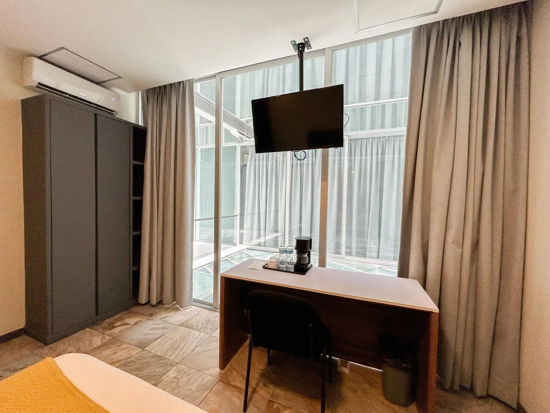 Habitación de hotel moderna con escritorio, televisor montado en la pared, armario, cortinas y gran ventanal con vistas.