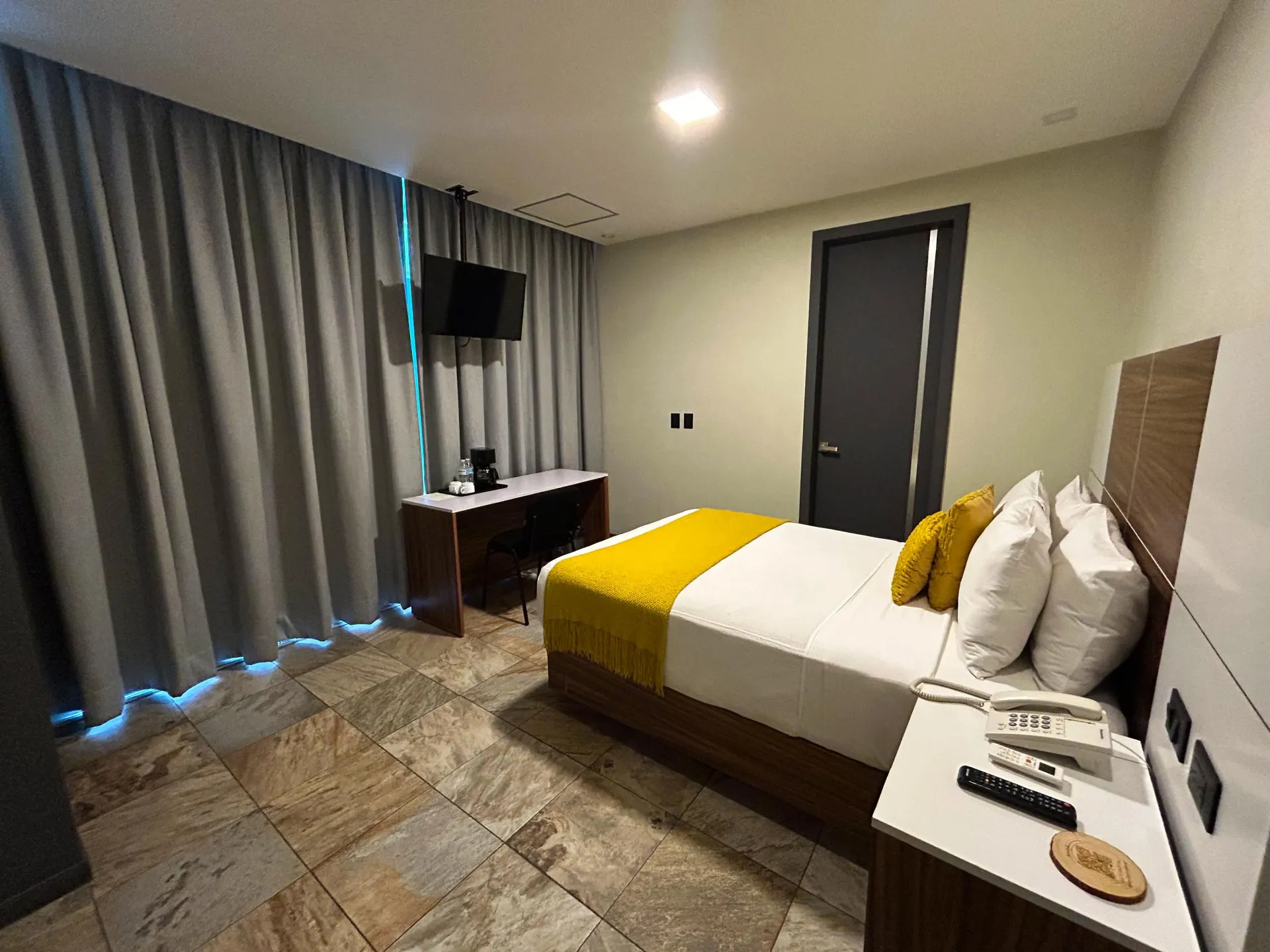 Habitación de hotel con dos camas, cortinas grises, TV, escritorio y ropa de cama con detalles en amarillo.
