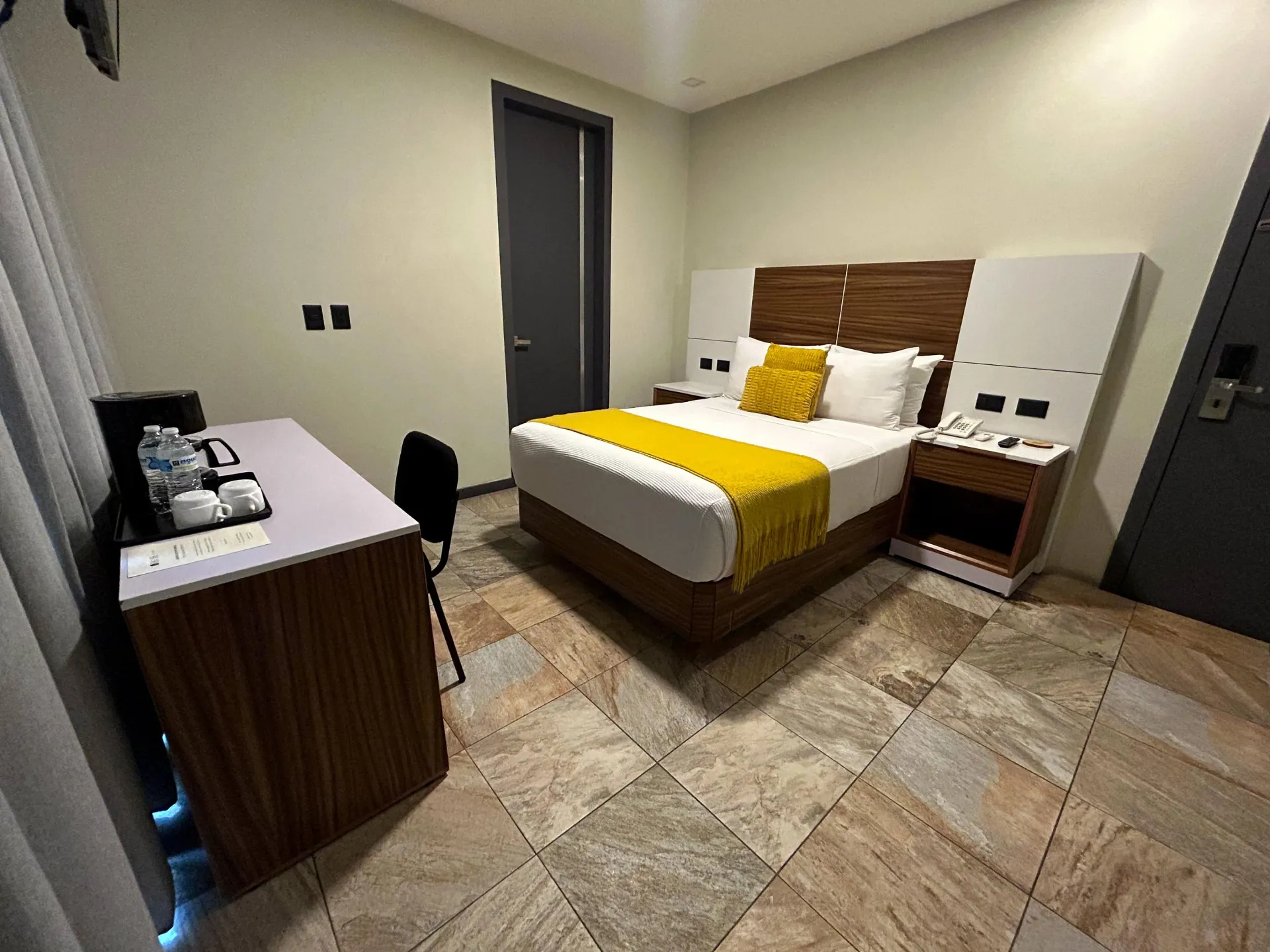 Habitación de hotel moderna con cama, escritorio, silla y suelo de baldosas en tonos neutros.