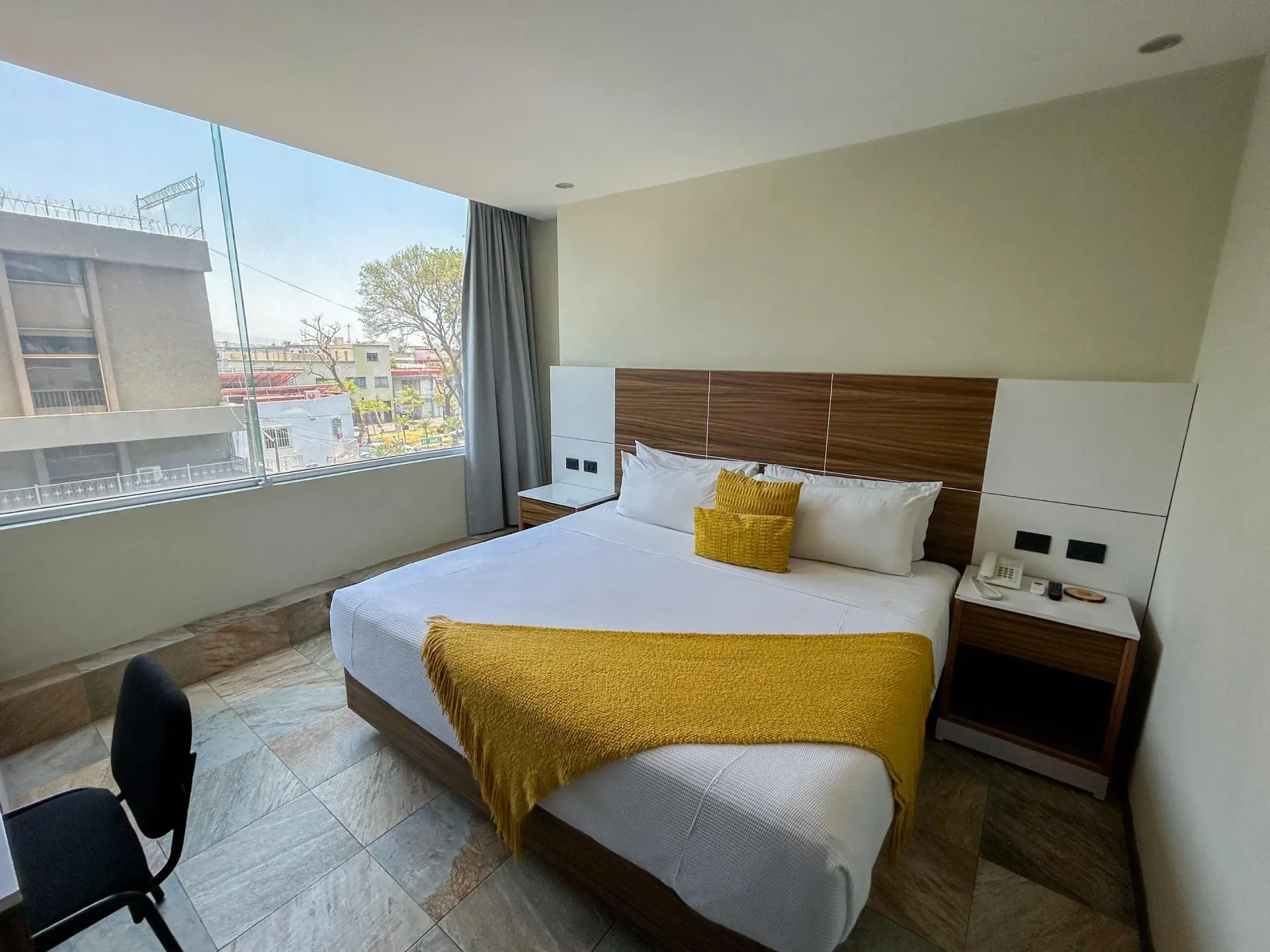 Habitación luminosa con una cama grande, una manta decorativa amarilla, una silla de escritorio y vistas a la ventana.