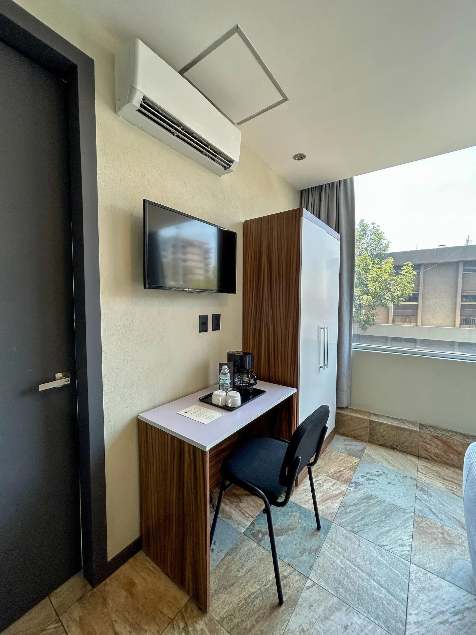 Pequeña habitación de hotel con escritorio, silla, televisor, cafetera, armario y balcón.