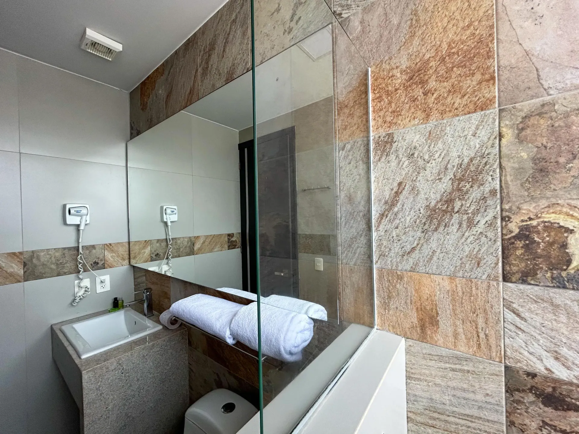 Baño moderno con paredes revestidas de piedra, mampara de ducha de cristal, lavabo y toallas blancas dobladas.
