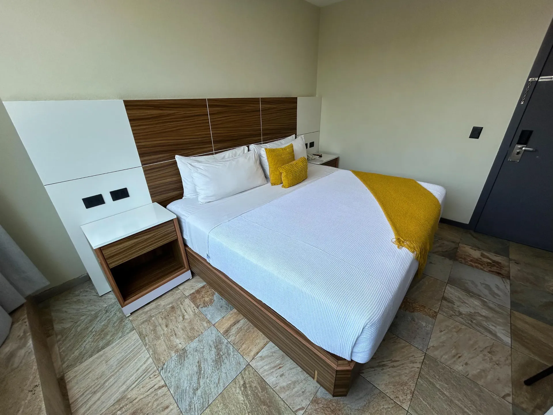 Habitación pequeña con cama, mesita de noche de madera, detalles en amarillo y suelo de baldosas.
