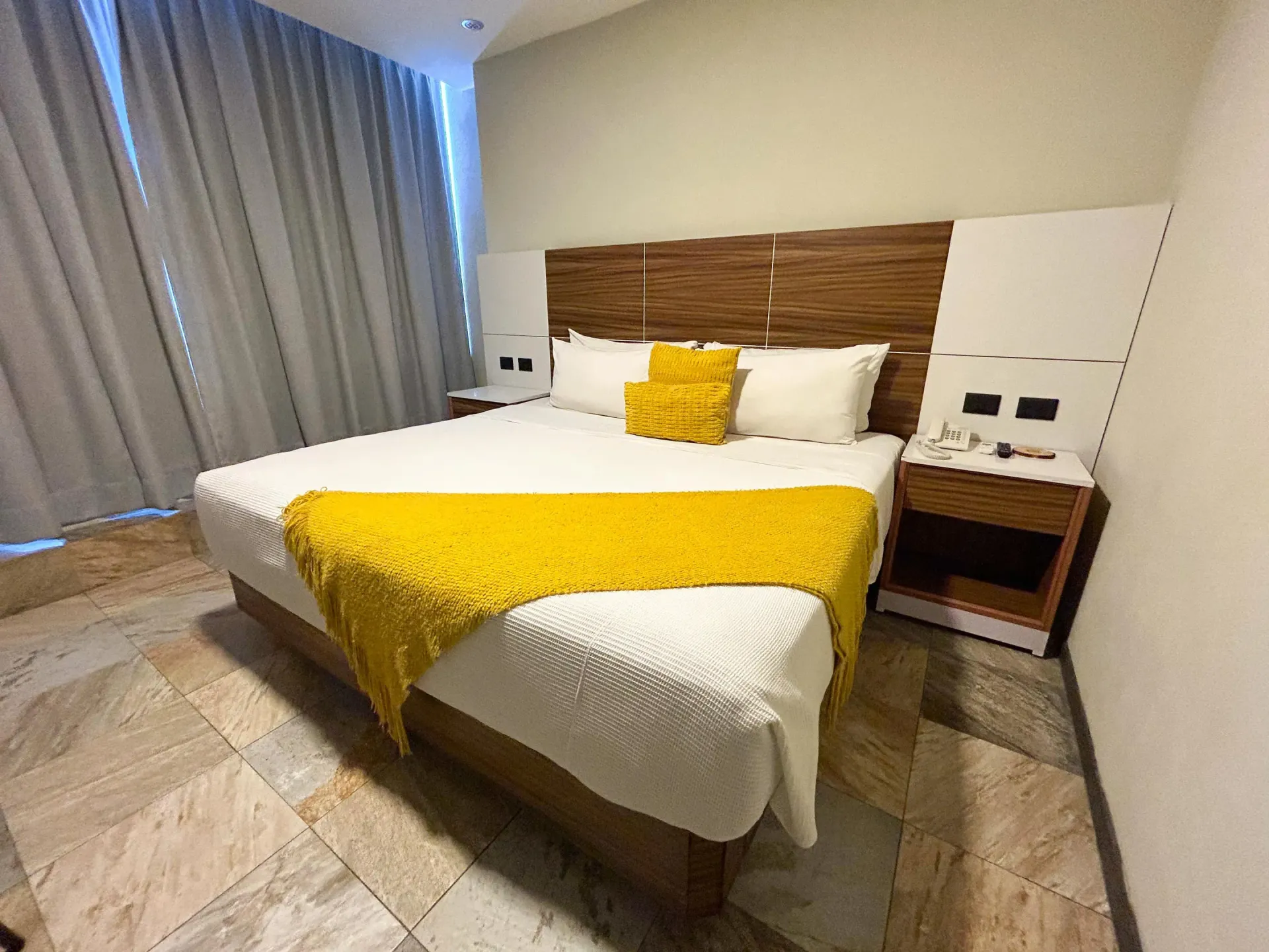 Habitación de hotel moderna con cama king size, manta amarilla, mesitas de noche y cortinas grandes.