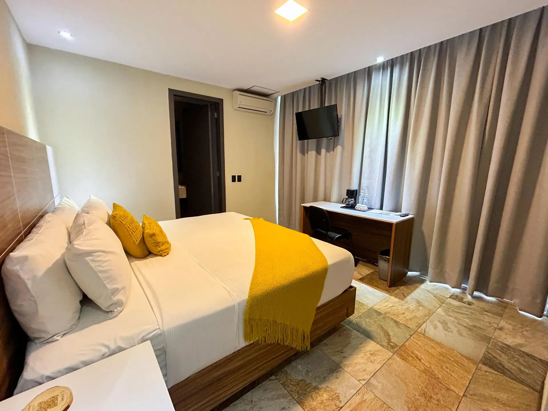 Dormitorio con cama, manta amarilla, televisor montado en la pared, escritorio y cortinas grises.