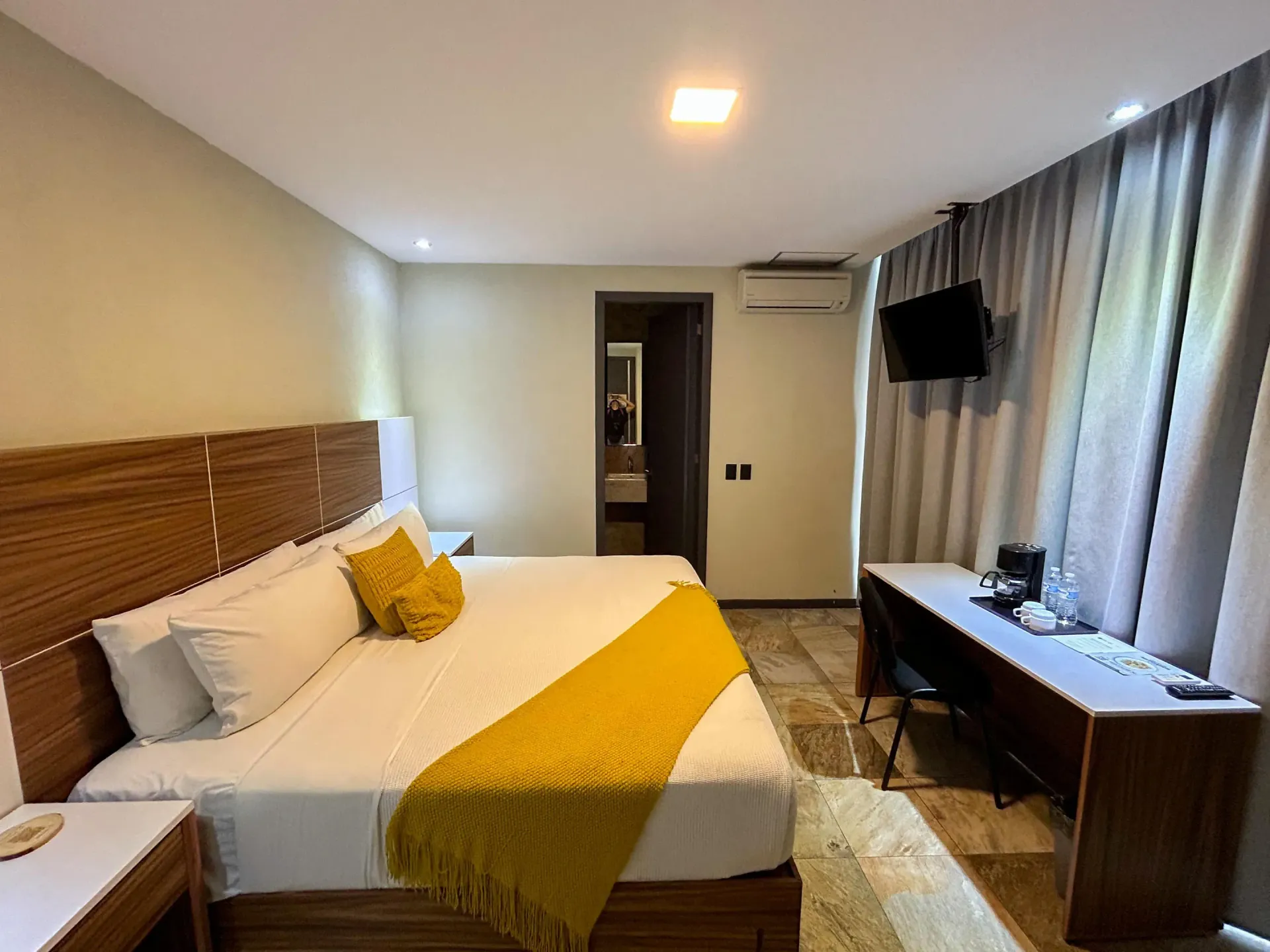 Habitación de hotel moderna con cama, manta amarilla, escritorio, televisor de pared y cortinas.