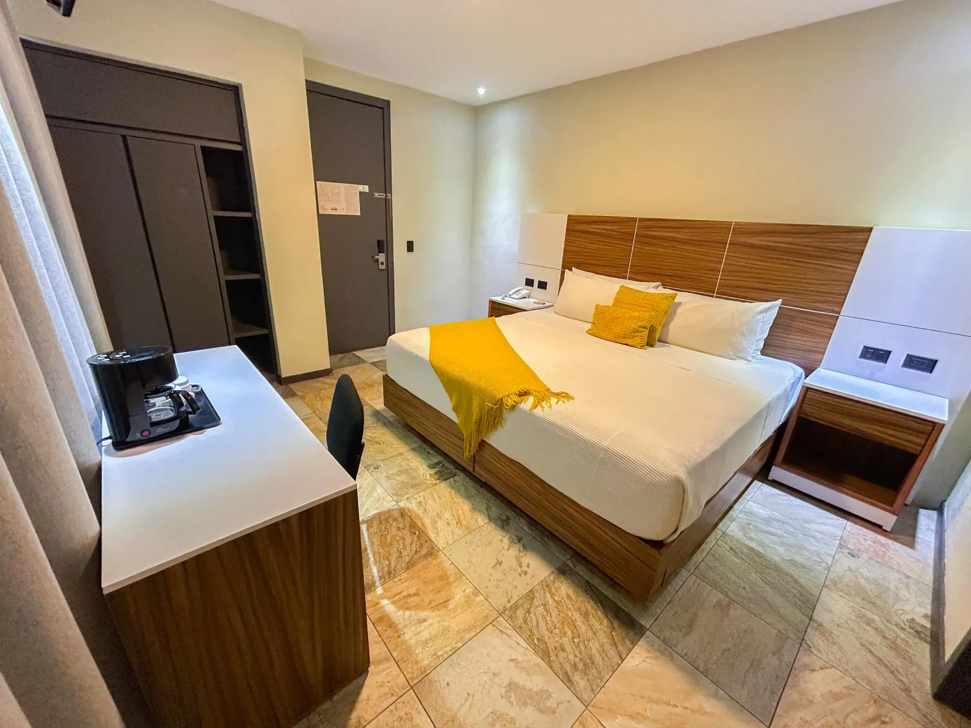 Dormitorio moderno con cama de madera, detalles en amarillo, armario y luminosa zona de estar junto a la ventana.