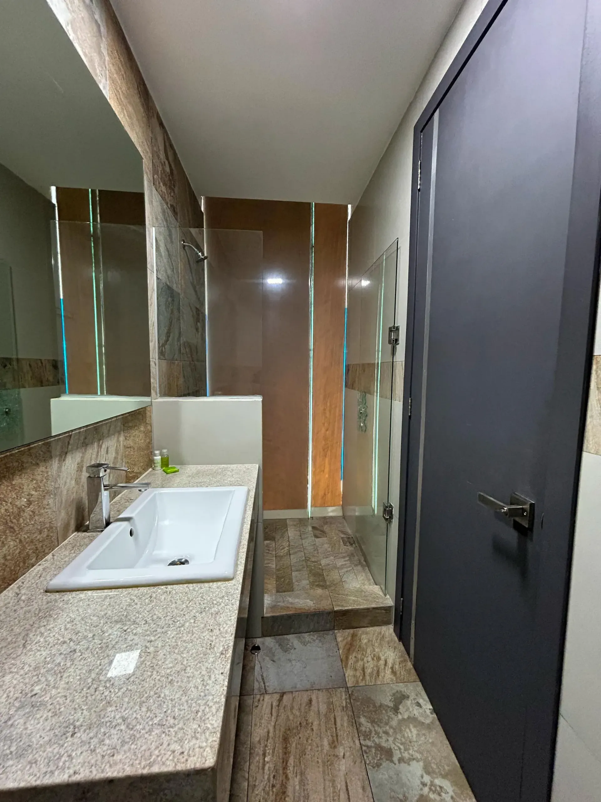 Baño moderno con lavabo de granito, espejo grande, ducha con azulejos marrones y puerta oscura cerrada.