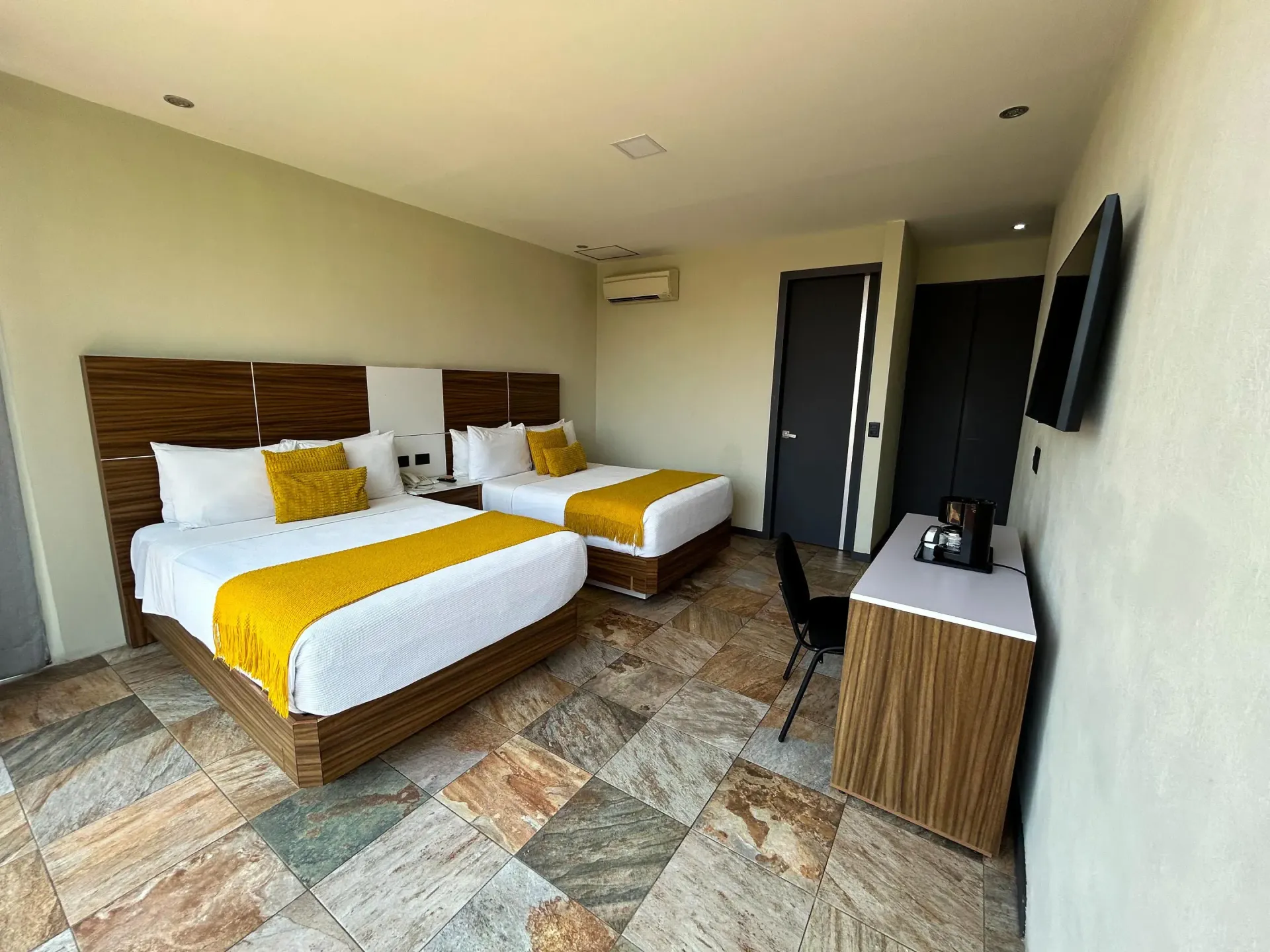Habitación de hotel con dos camas, detalles en amarillo, suelo de baldosas y televisor de pared.