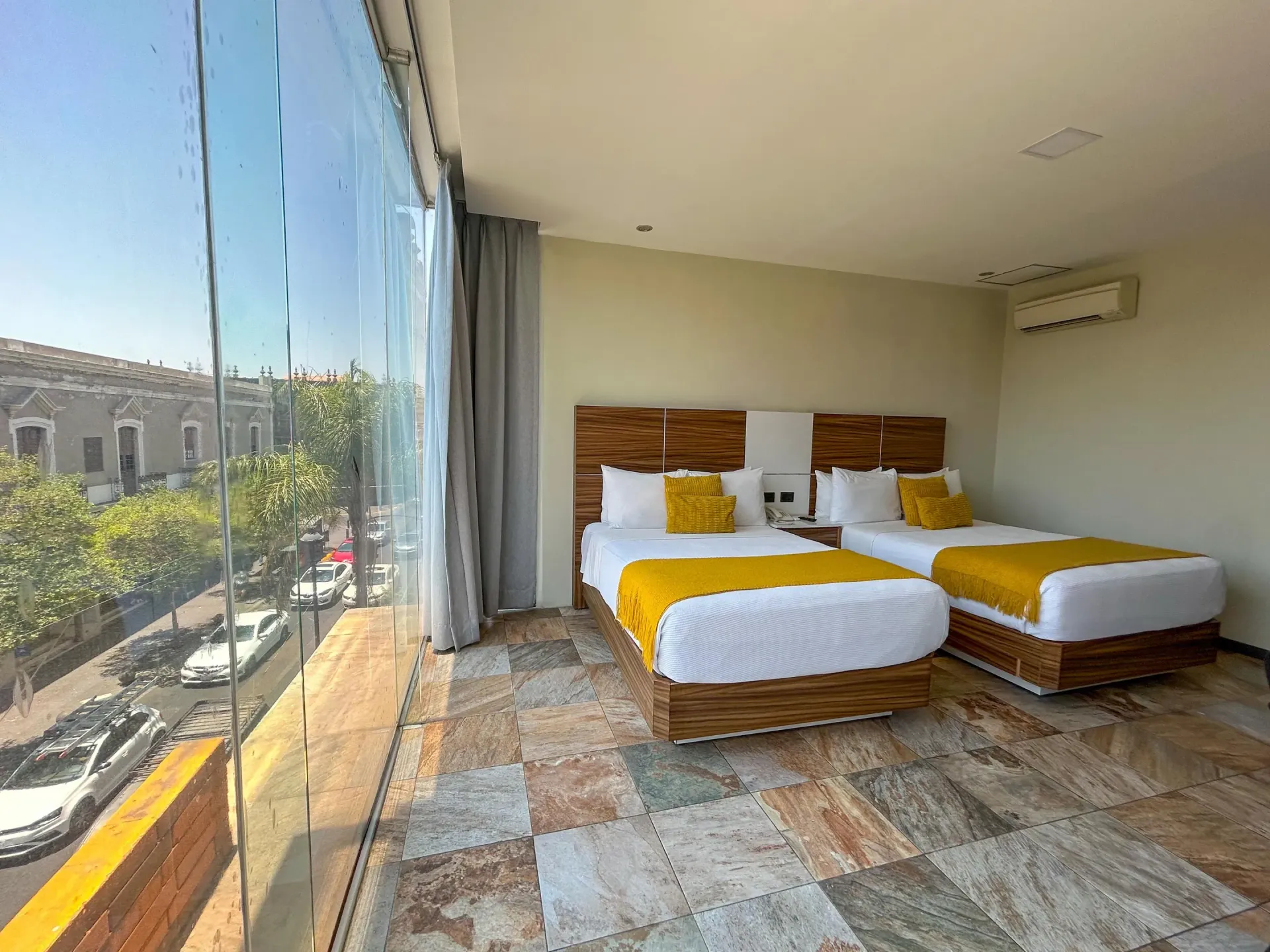 Habitación de hotel luminosa con dos camas individuales, detalles en amarillo y una gran ventana con vistas a la calle.