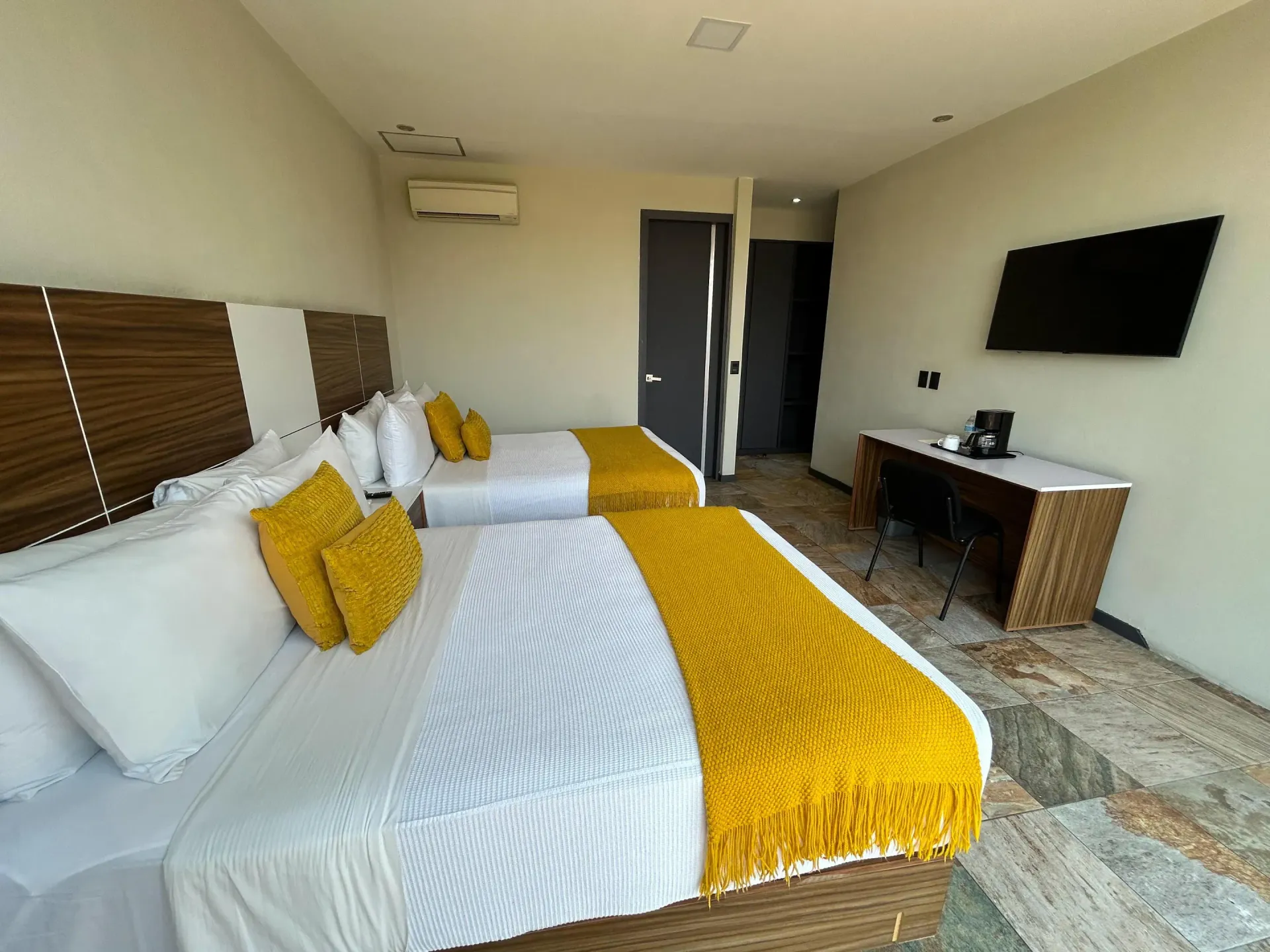 Habitación de hotel con dos camas individuales, detalles en amarillo, escritorio, televisor de pared y puerta al balcón.