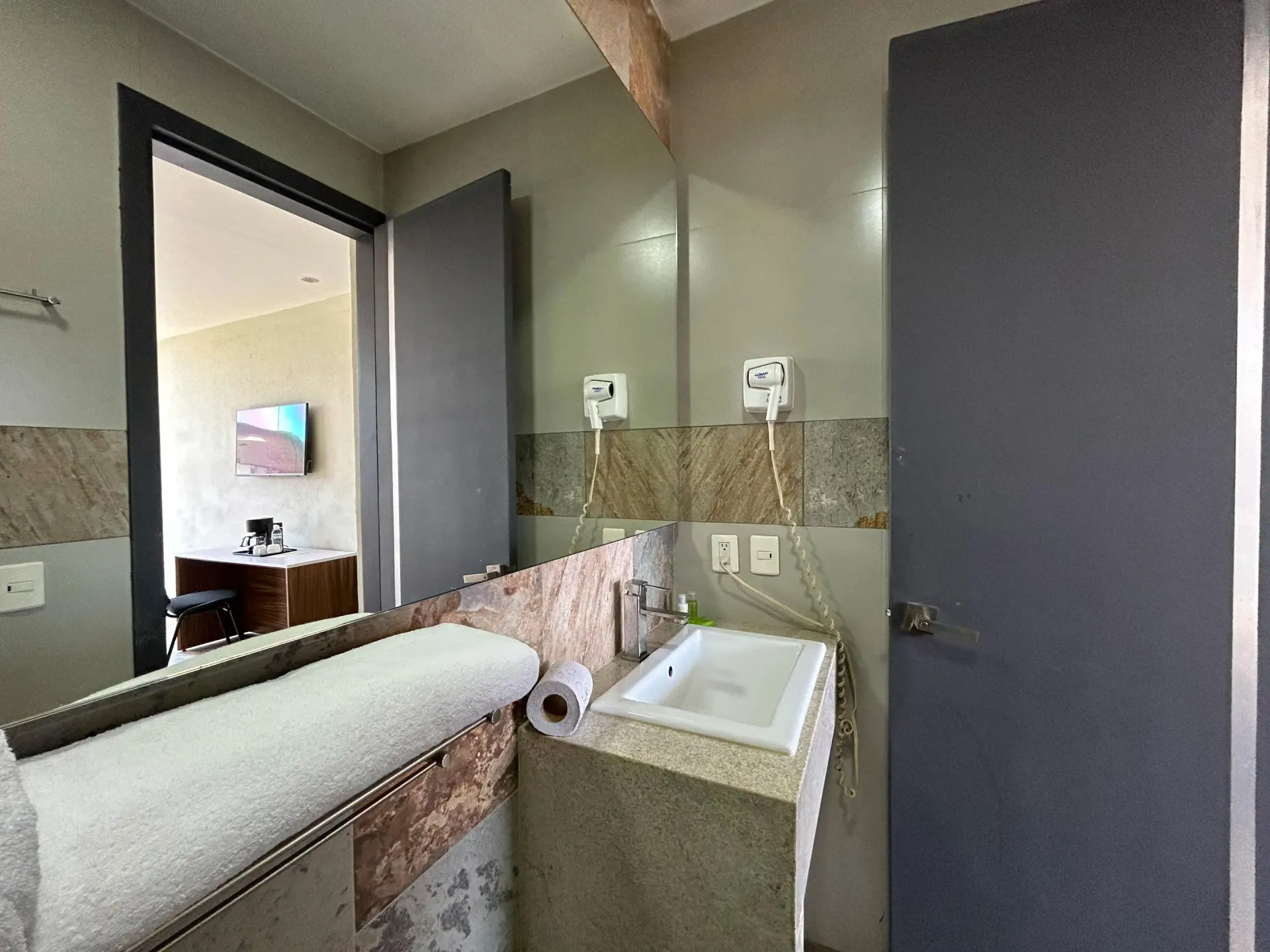 Baño con lavabo, espejo, dispensador de toallas de papel y puertas de color gris oscuro en una habitación alicatada.