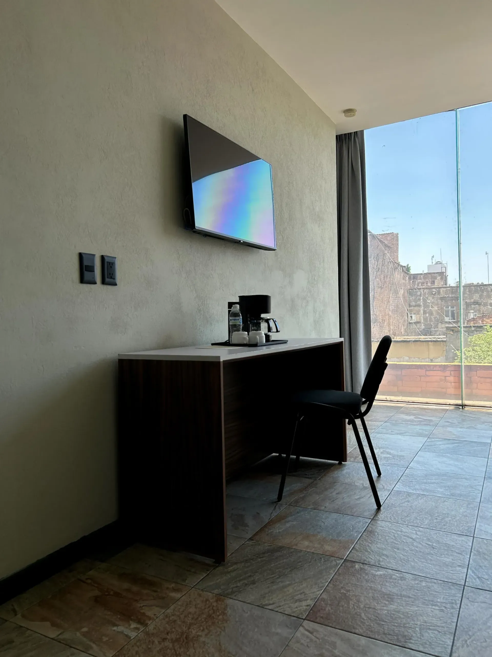 Habitación moderna con televisor de pared, escritorio y silla junto a una ventana con vistas a la ciudad.