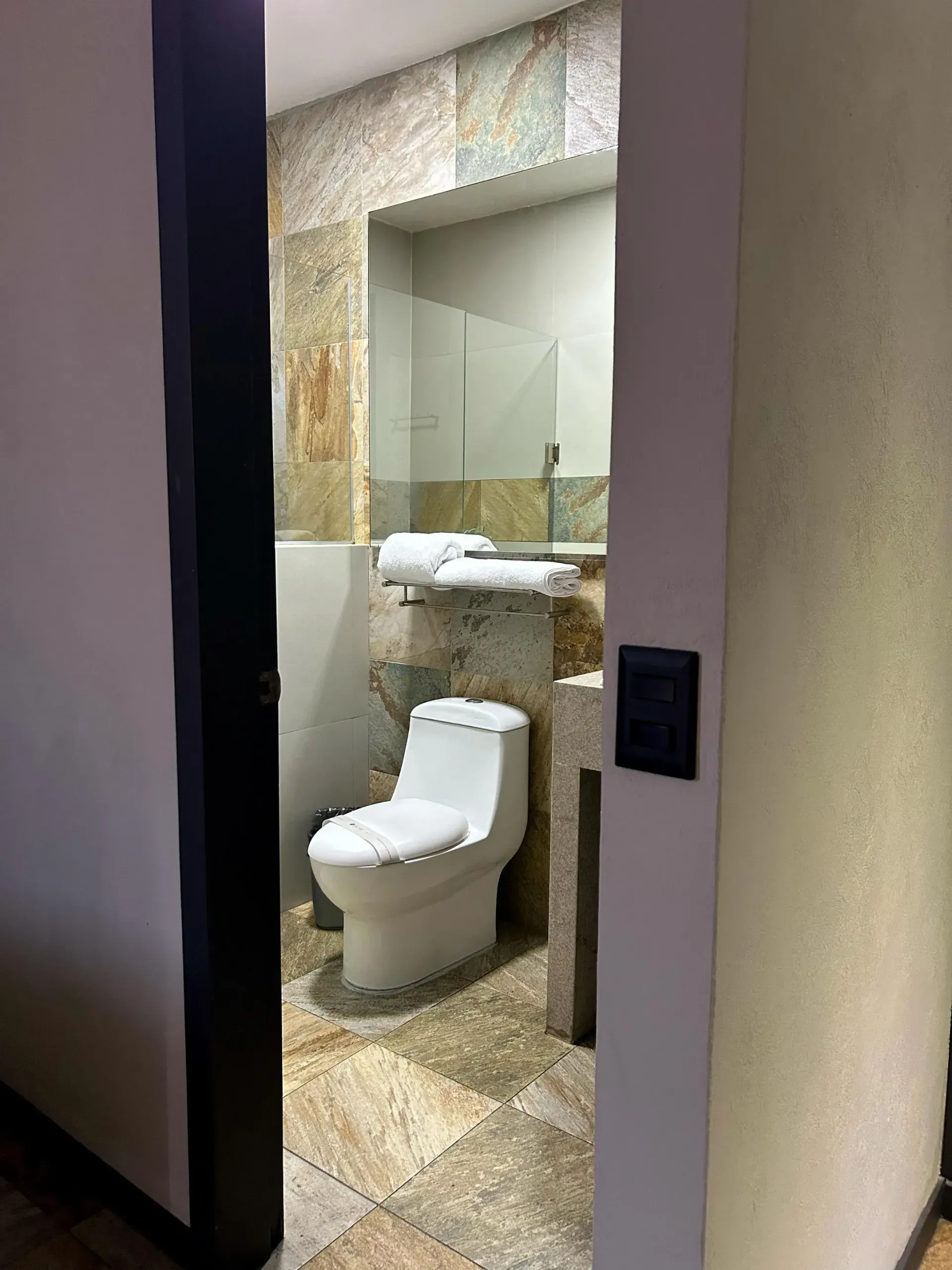 Pequeño aseo con inodoro y lavabo, visible a través de una puerta, con azulejos de piedra beige.