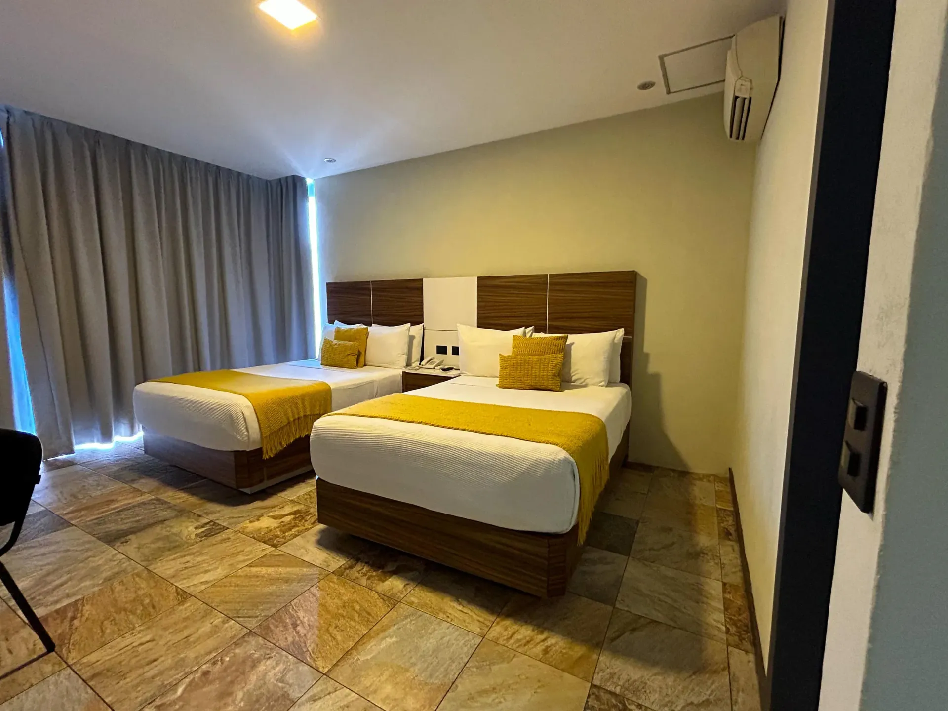 Habitación de hotel con dos camas bien hechas, cortinas, iluminación cálida y suelo de baldosas.