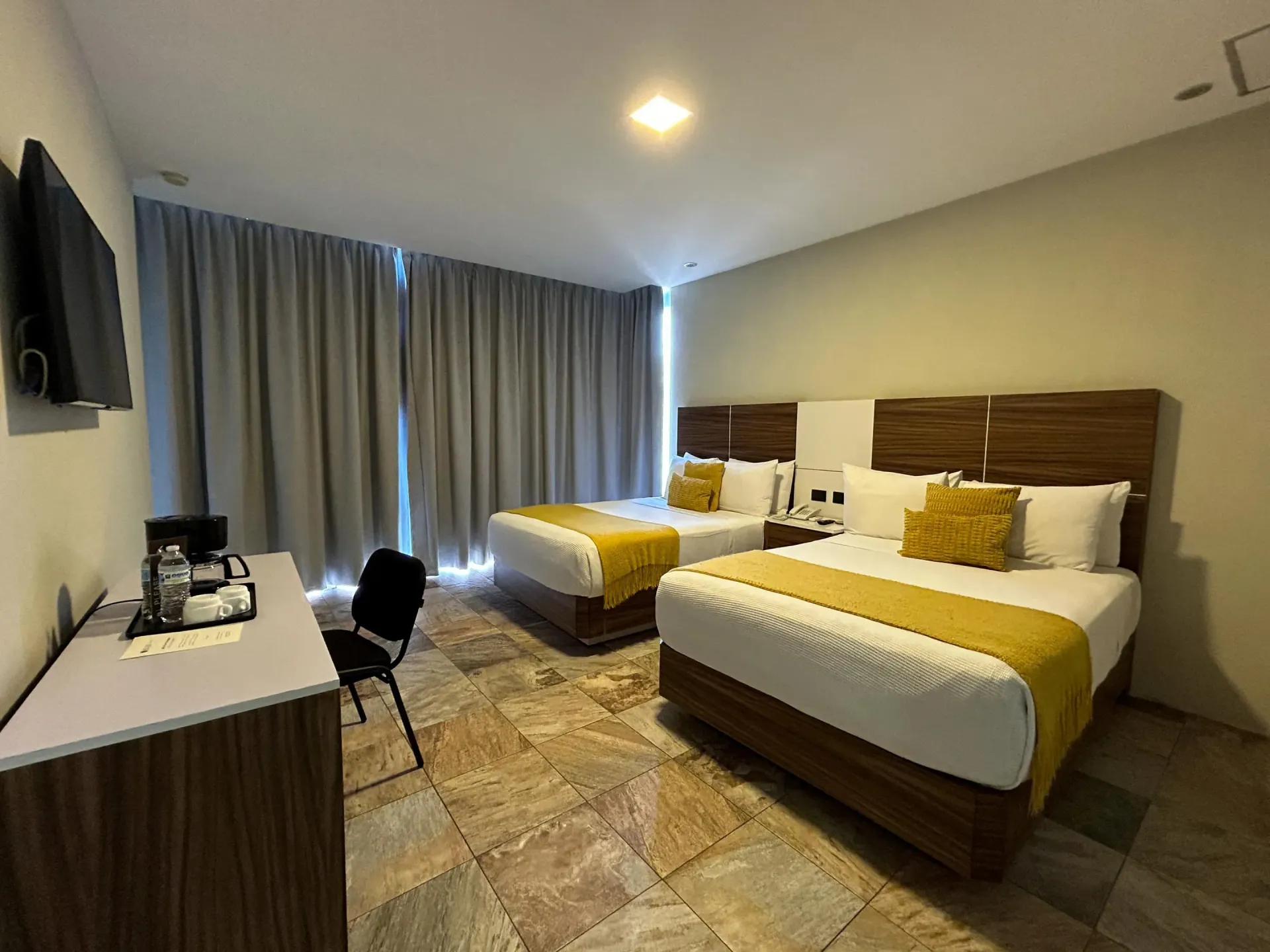 Habitación de hotel con dos camas, cortinas grises, un escritorio y detalles en amarillo cálido.