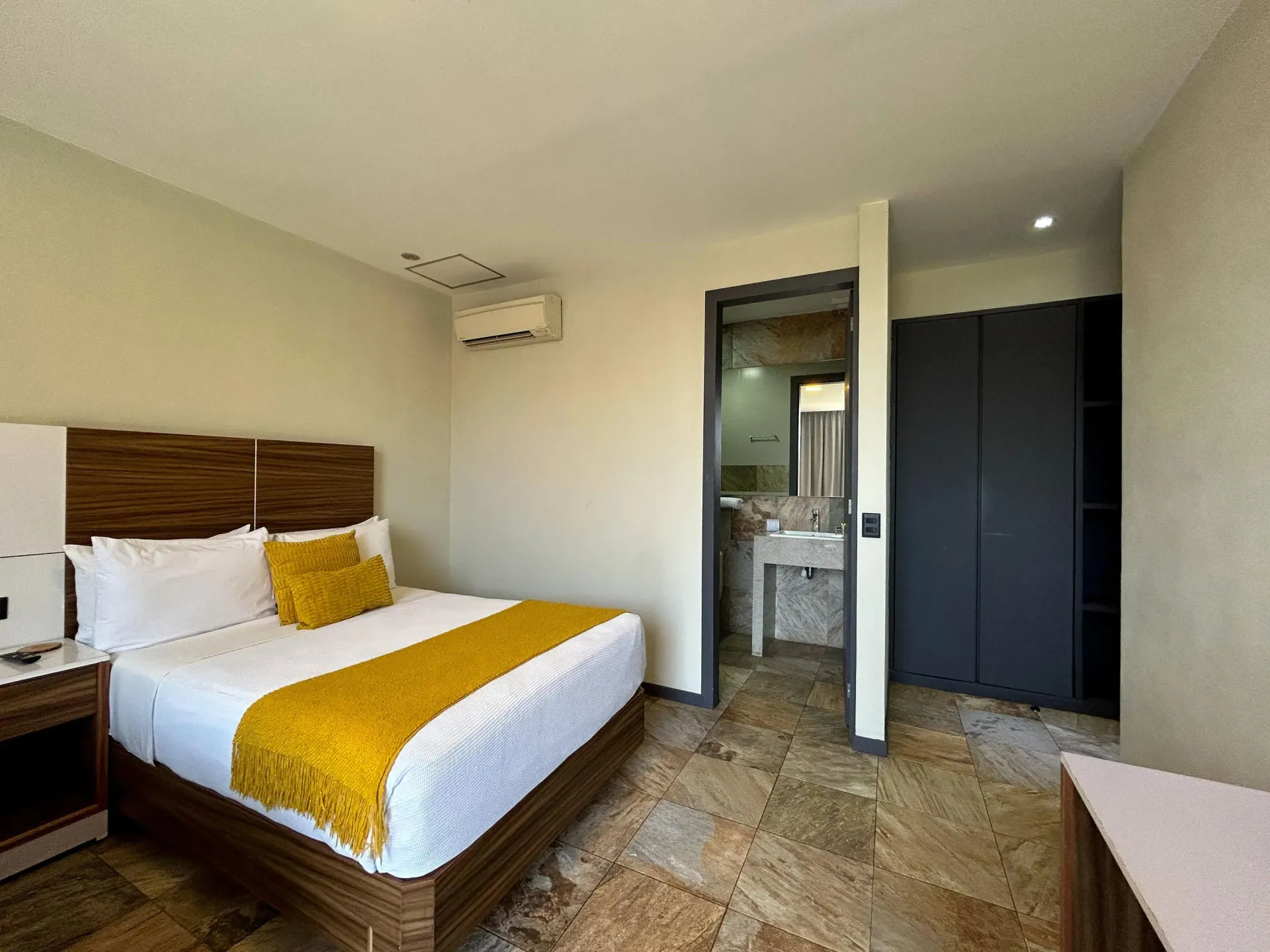 Habitación de hotel moderna con cama, manta amarilla, suelo de baldosas y baño visible a través de una puerta abierta.