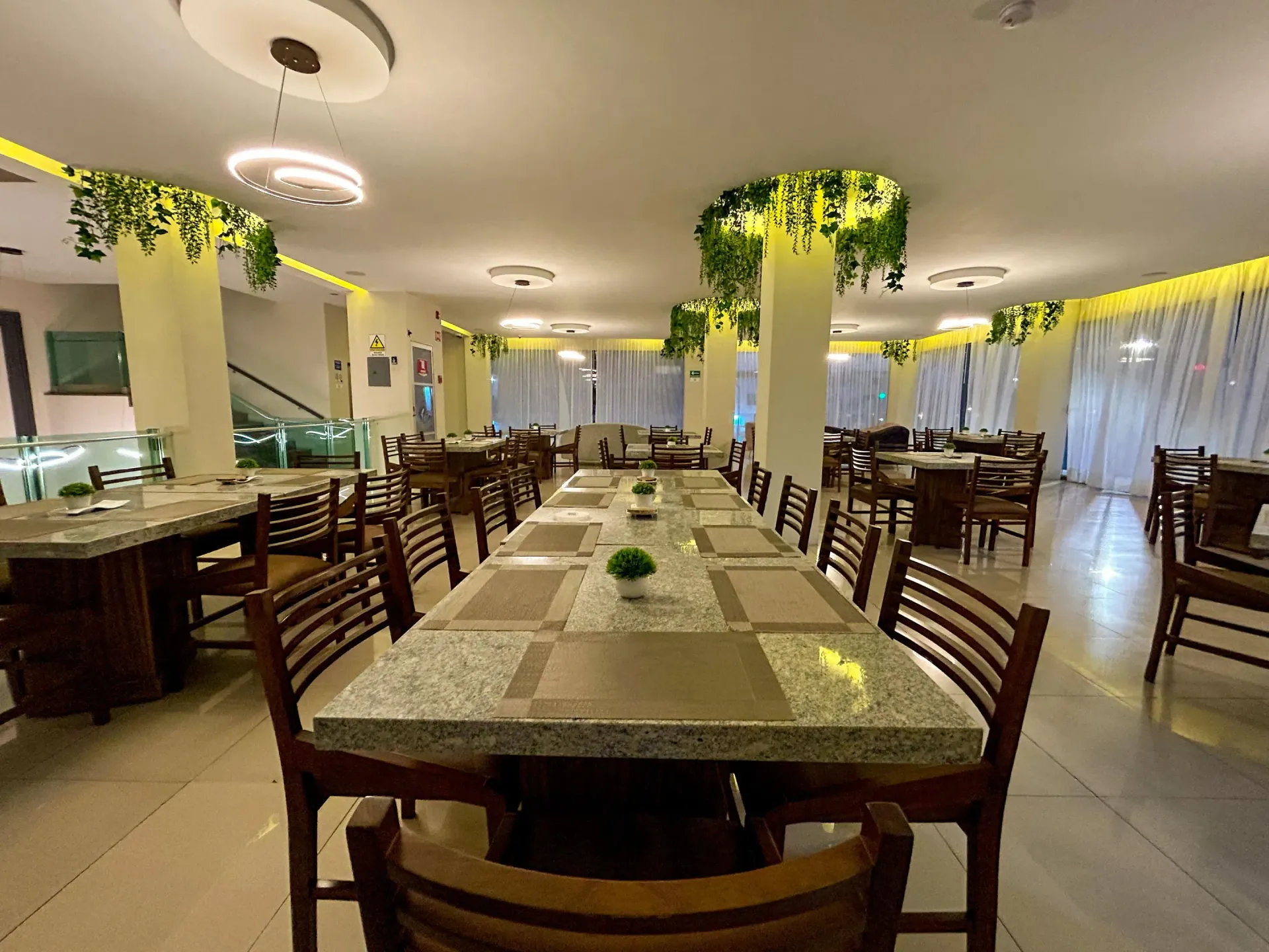 Elegante salón de banquetes con largas mesas de comedor, sillas de madera y plantas colgantes bajo cálidas luces de techo.