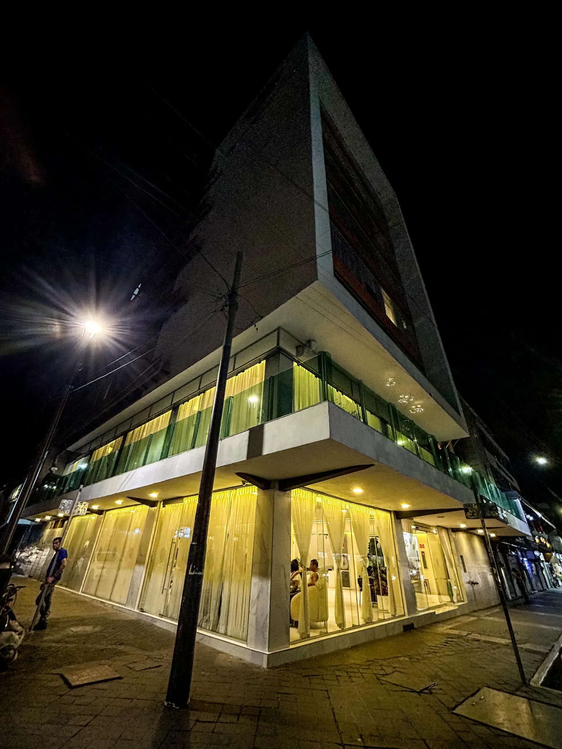 Vista nocturna de un moderno edificio de esquina con fachada de cristal y una brillante iluminación interior amarilla.