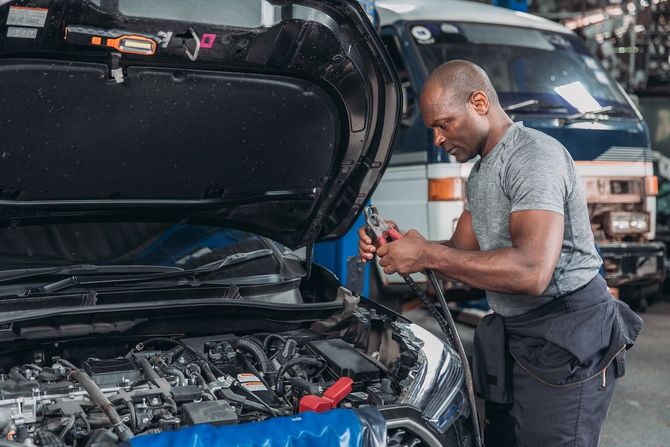 Mechanic working under an open car hood in a garage, holding a tool near the engine.