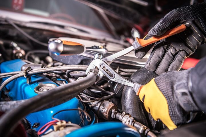 Mechanic working under a car hood, tightening engine parts with a wrench and wearing black-and-yellow gloves