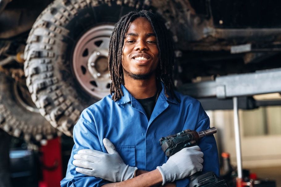 Mechanic in blue coveralls standing with arms crossed under a raised vehicle in a garage