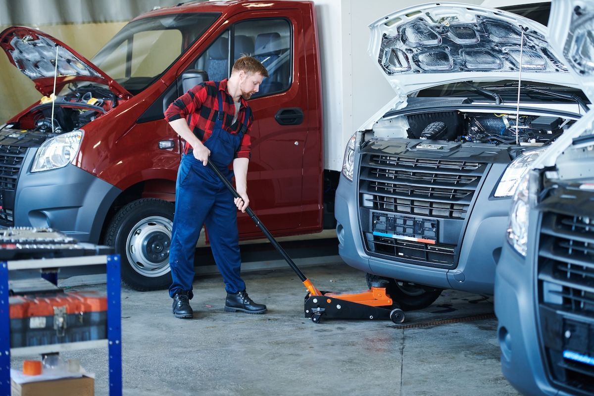 Mechanic using a floor jack beside a red van in an auto repair shop with cars open for service