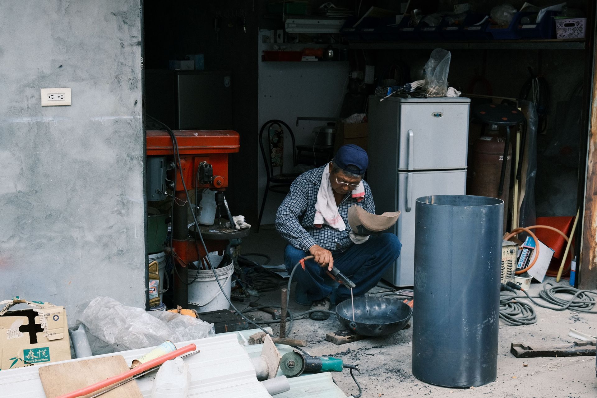 Person crouching in a cluttered garage, working on a motorcycle amid tools, boxes, and a refrigerator