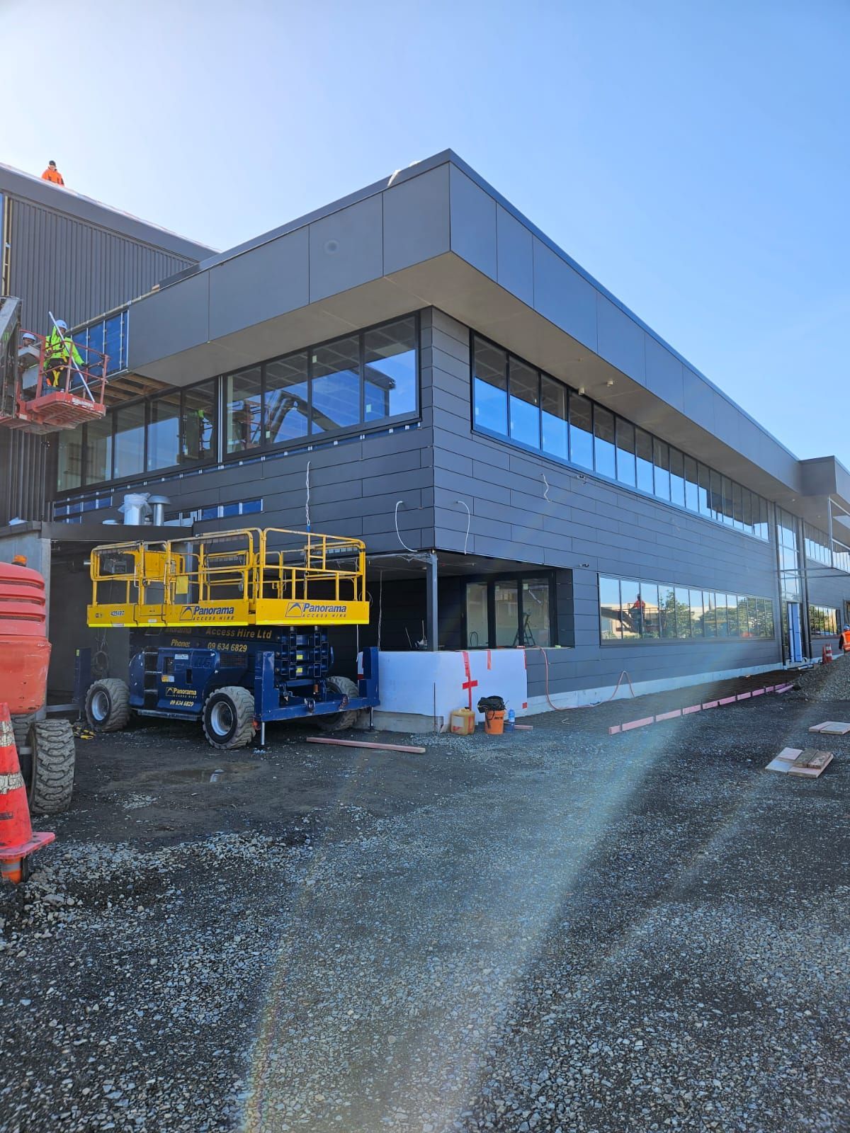 Modern building under construction; workers in lift, blue lift machinery on gravel, blue sky.