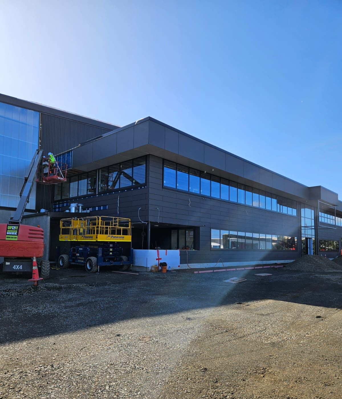 Building construction site with a worker in a lift. Gray facade, blue sky, and gravel ground.