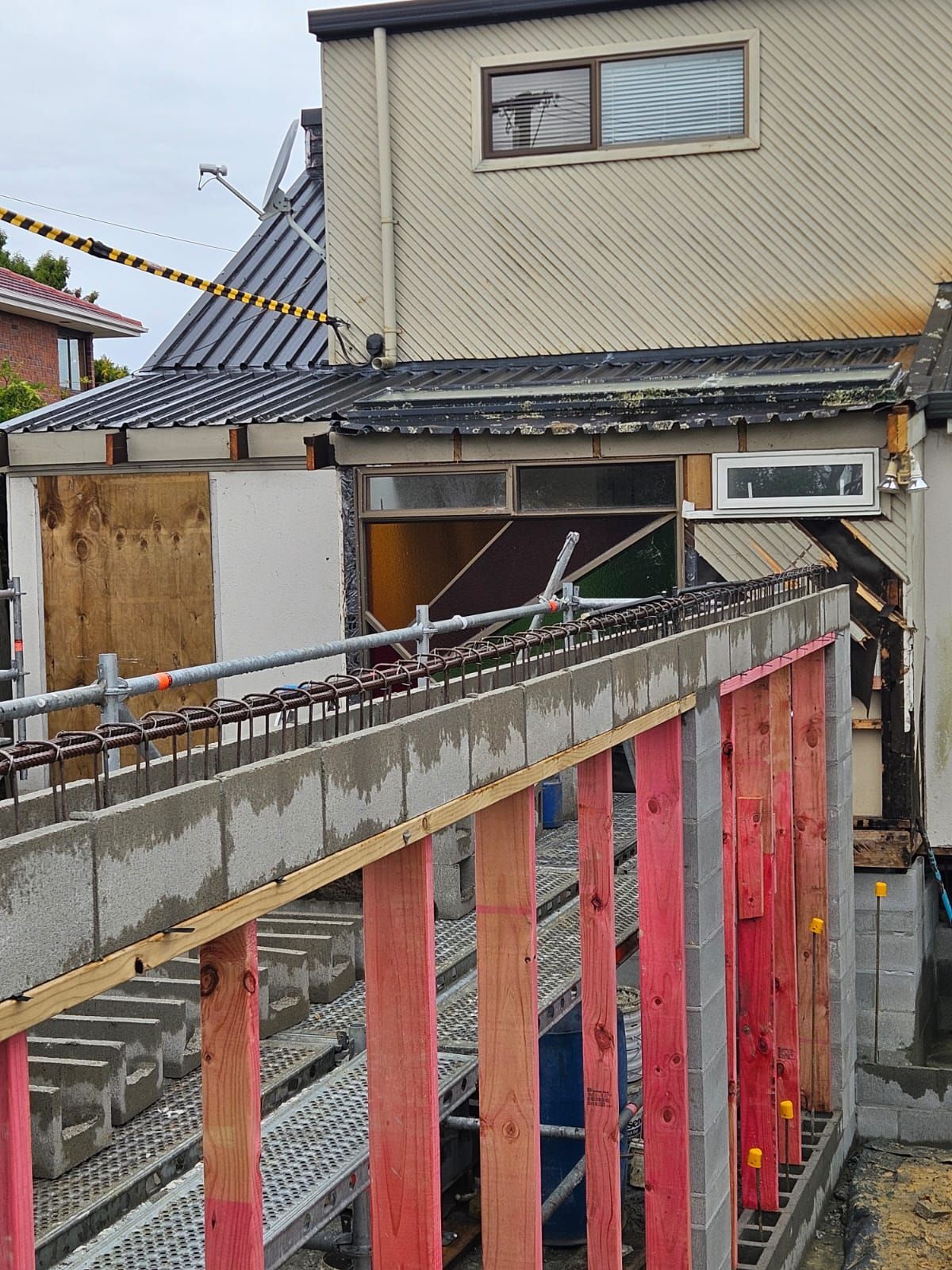 Construction site: partially built concrete wall with rebar, next to a two-story building.