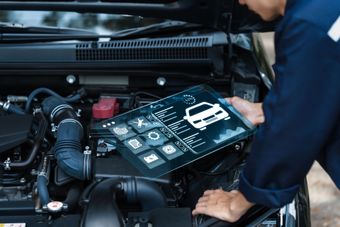 Person inspecting car engine with a diagnostic tablet in the open hood area | Cain And Sons' Automotive