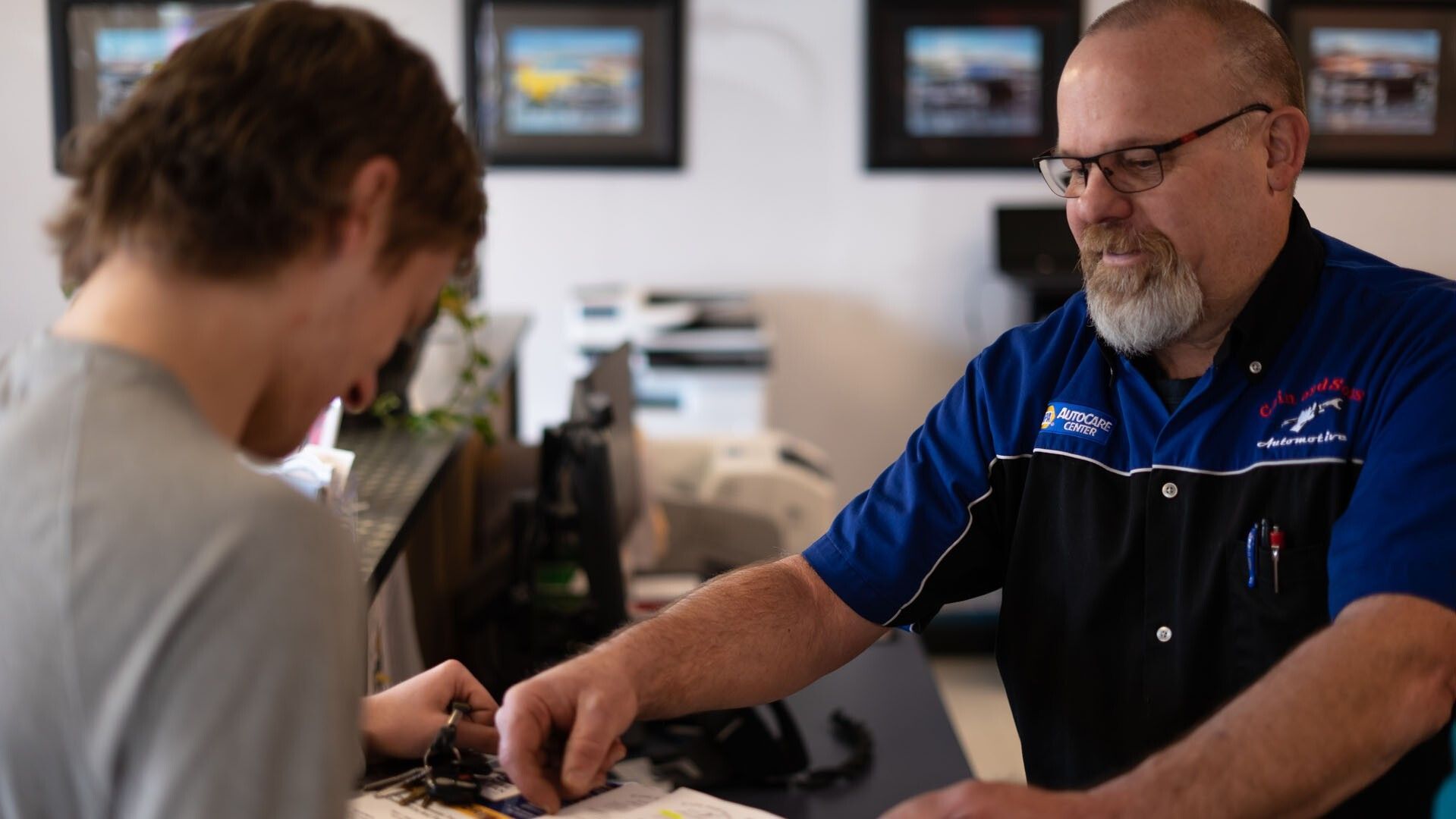 Two men seated at a table, one handing over an item in a bright office or shop setting. | Cain And Sons' Automotive