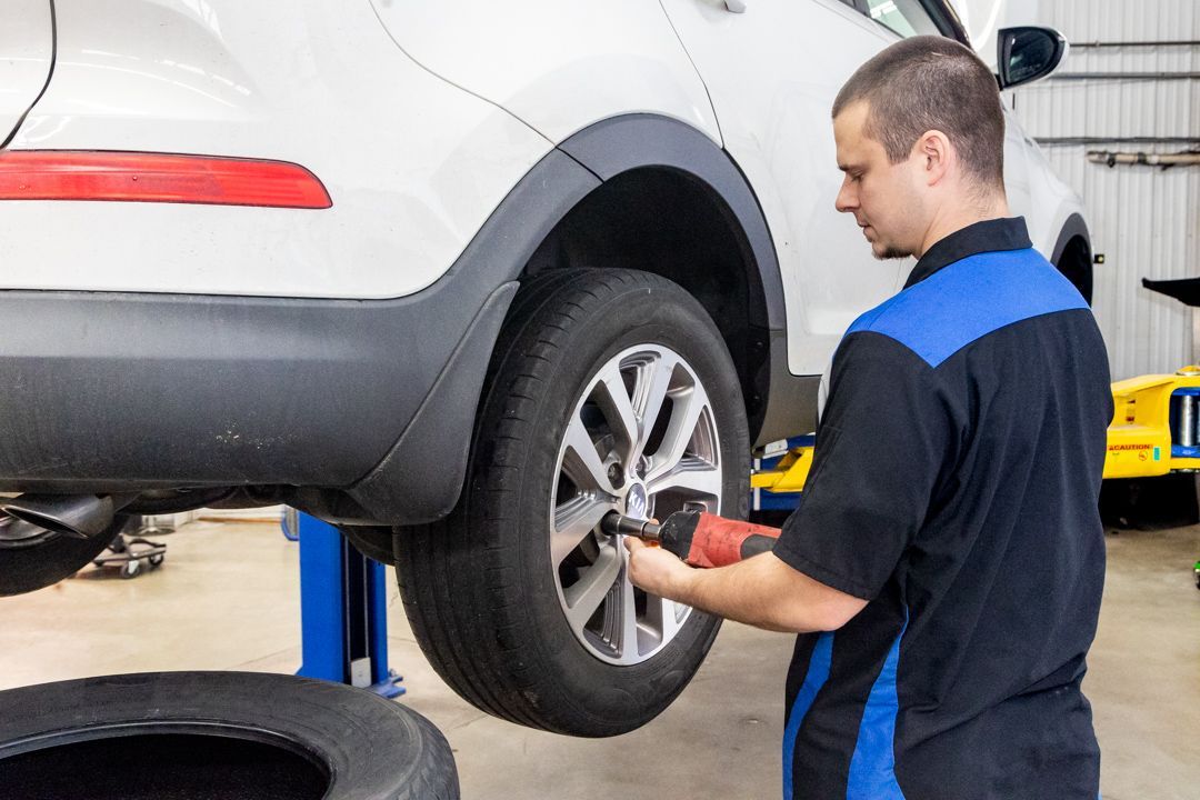 Mechanic working on a silver car’s rear wheel in an auto shop with a blue lift | Cain And Sons' Automotive