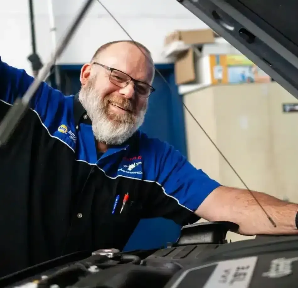 Mechanic in a blue uniform smiling while working under an open car hood in a garage | Cain And Sons' Automotive