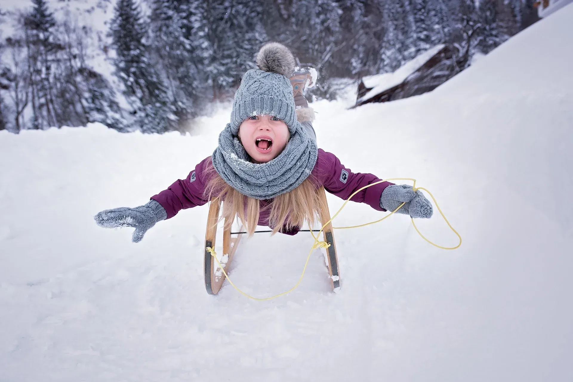 Child sledding down a snowy hill, wearing a hat, scarf, and mittens, with an open-mouthed expression of joy.