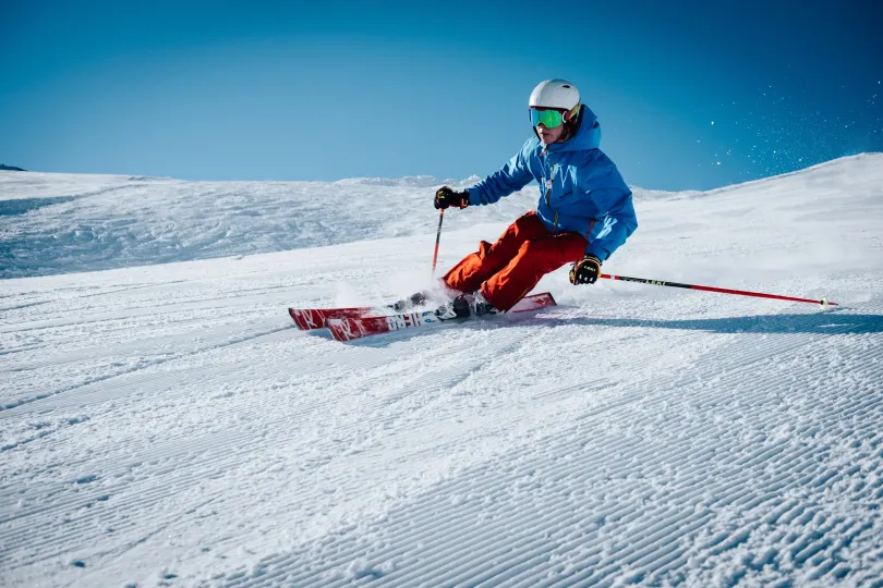 Skier in blue jacket and orange pants carves down a snowy slope on a bright, sunny day.