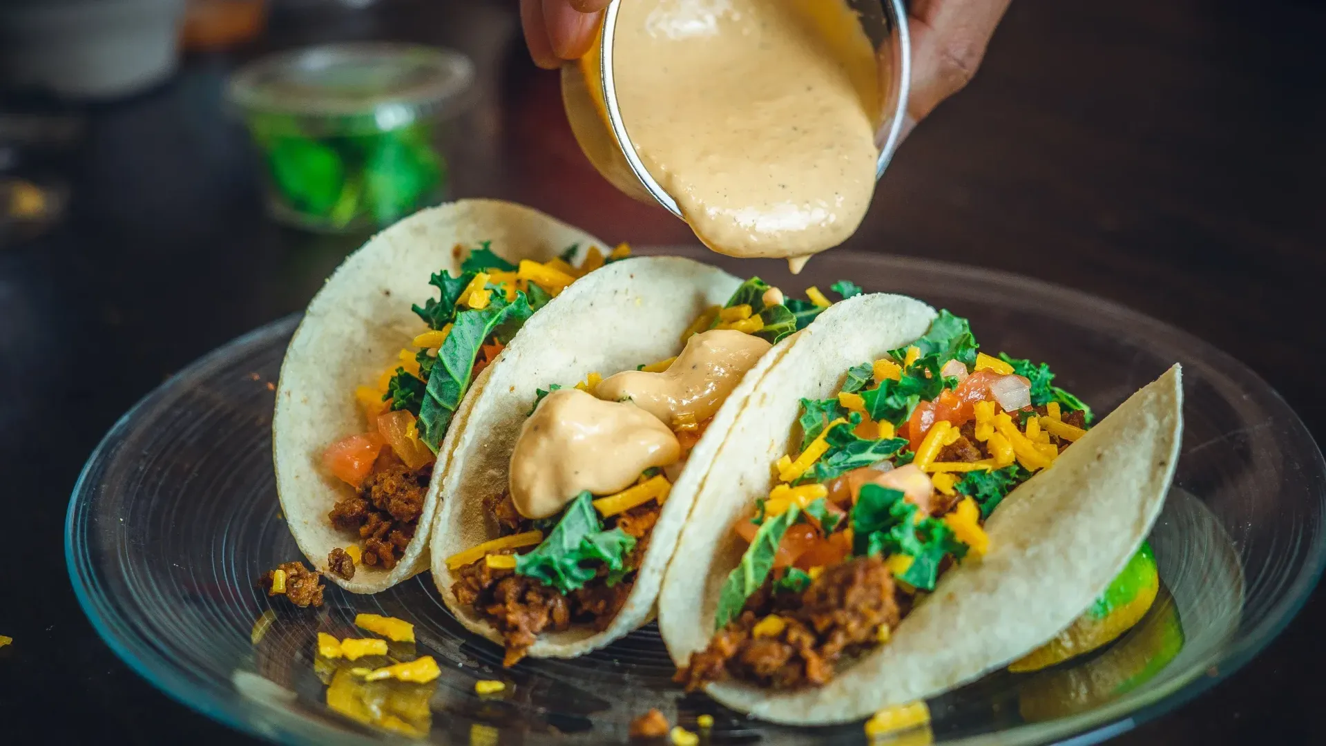 Tacos on a plate being drizzled with sauce. Green kale, cheese, and meat filling in each.