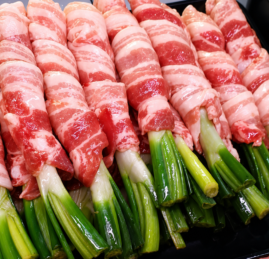 Cipollotti verdi crudi avvolti nella pancetta e disposti in file, pronti per la cottura.