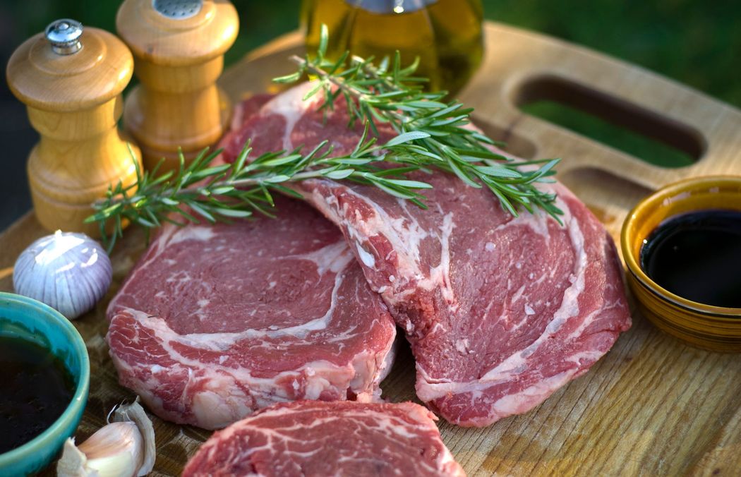 Bistecche di manzo crude con rosmarino su un tagliere, con macinapepe e ciotoline nelle vicinanze.