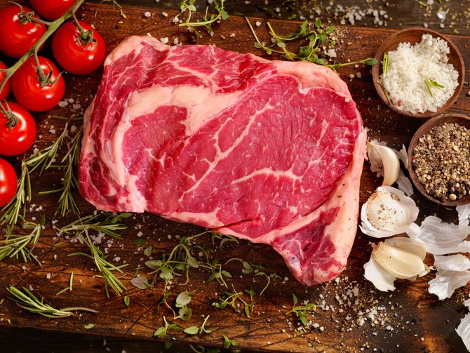 Bistecca cruda marmorizzata su un tagliere di legno con pomodorini, erbe aromatiche, aglio, sale e grani di pepe.
