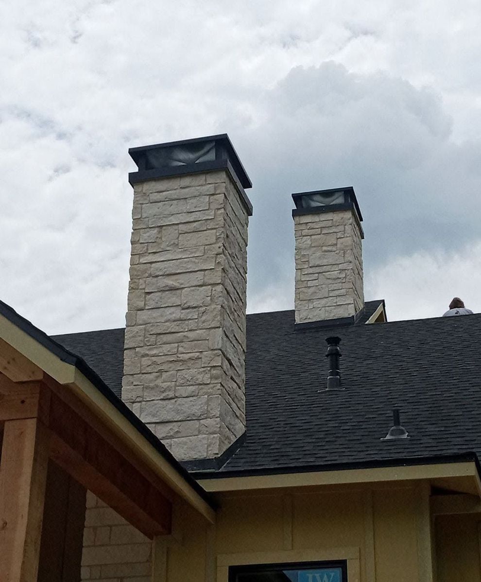 Two stone chimneys on a dark shingled roof under cloudy skies
