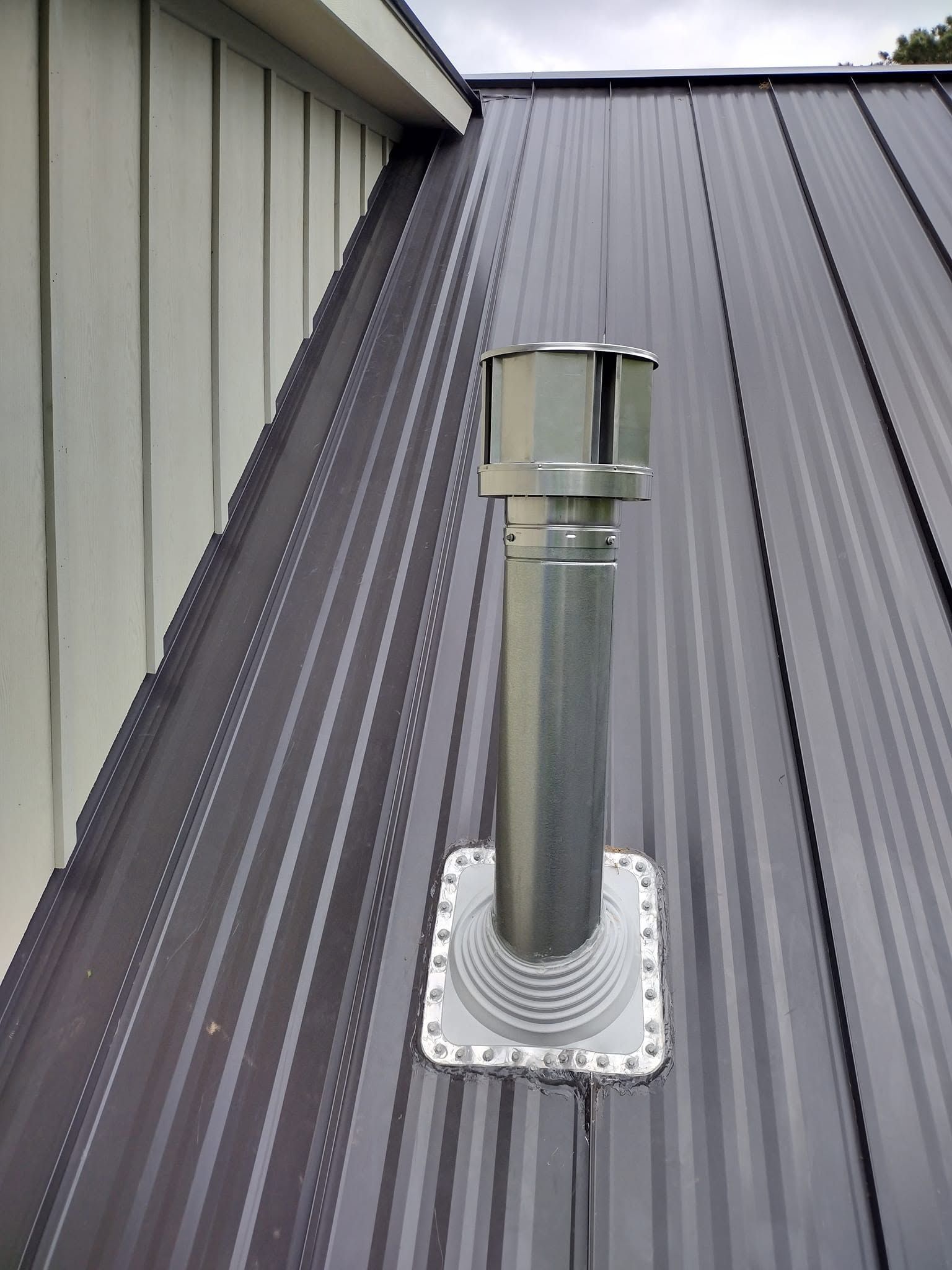 Metal roof vent pipe with cap on a gray standing-seam roof next to a wall