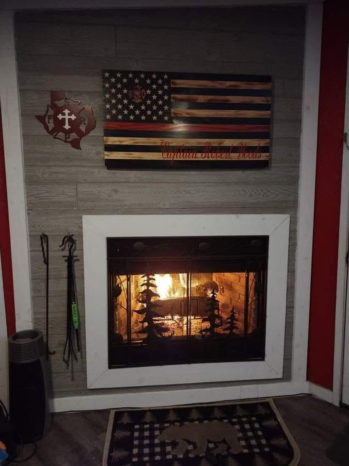 Cozy fireplace with white mantel, rustic American flag decor, and hanging tools on a gray stone wall.