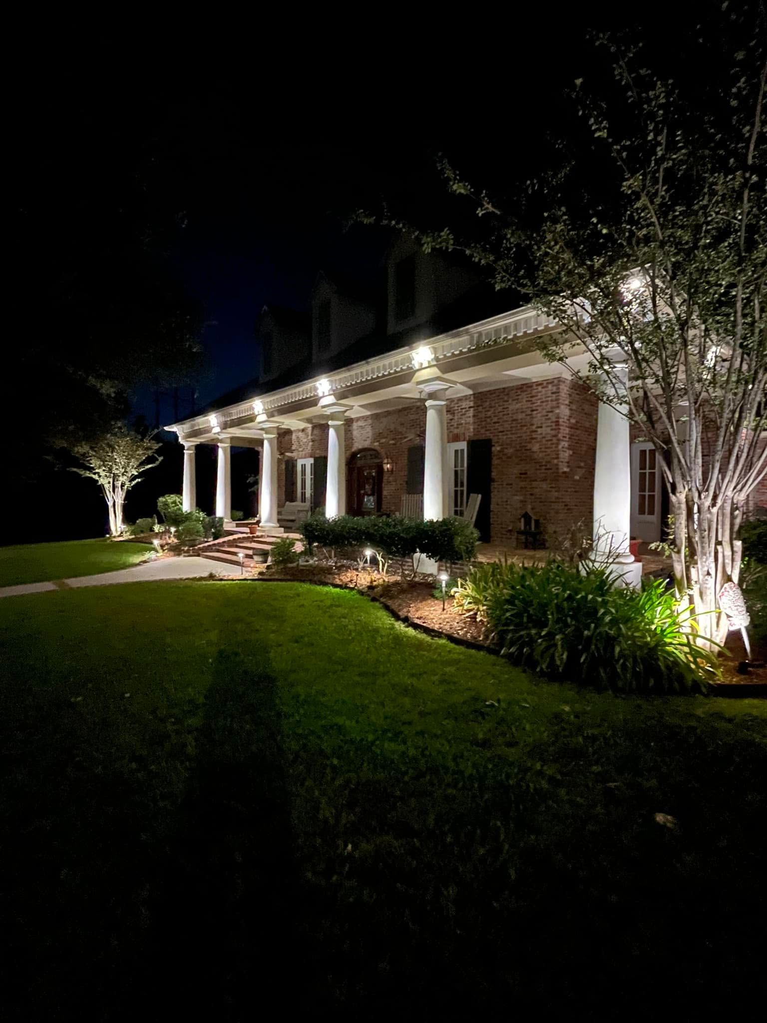 Well-lit brick house at night with a porch, landscaped lawn, and trees lit by exterior lights