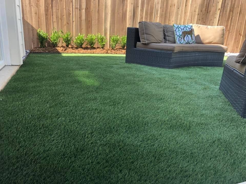 Backyard patio with artificial grass, potted plants, and a dark outdoor sofa with cushions.