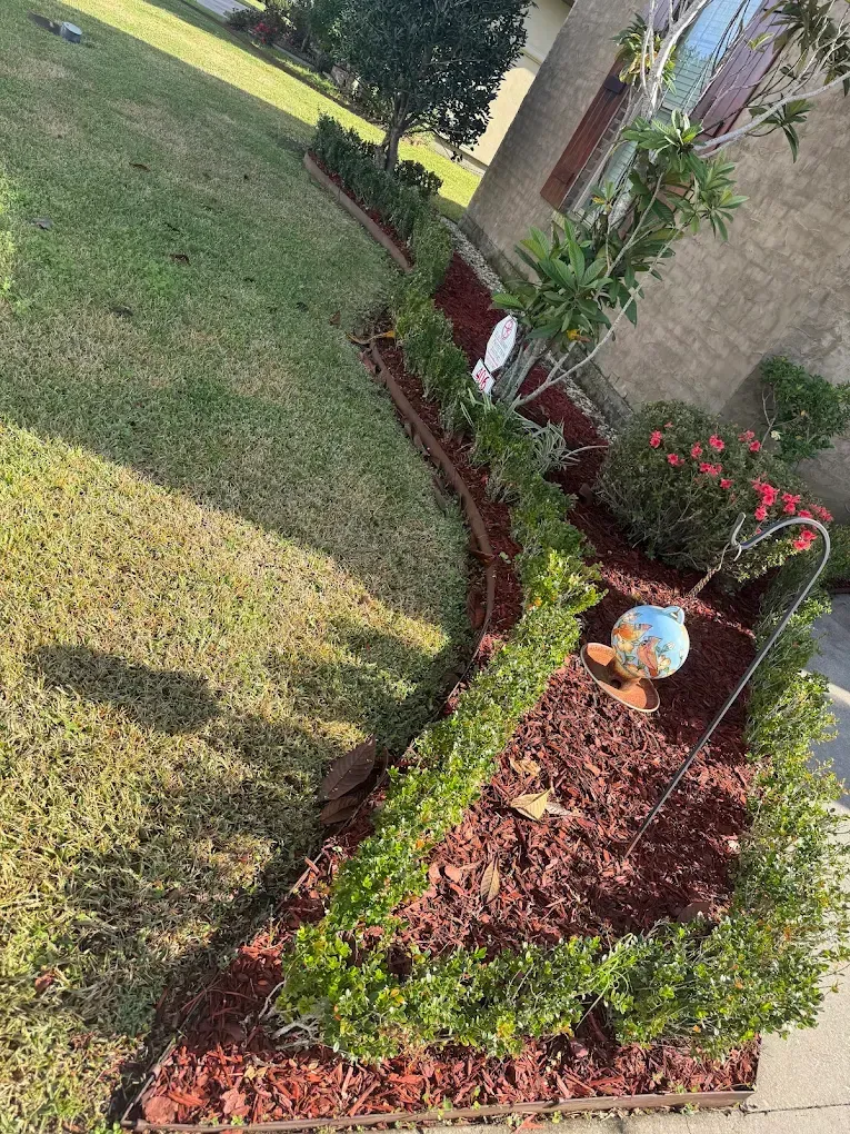 Curved flower bed with red mulch, green edging, and a small garden ornament beside a lawn and building.