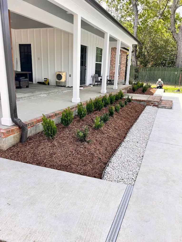 Covered porch with white columns, mulched planting bed, and a concrete walkway beside a house.