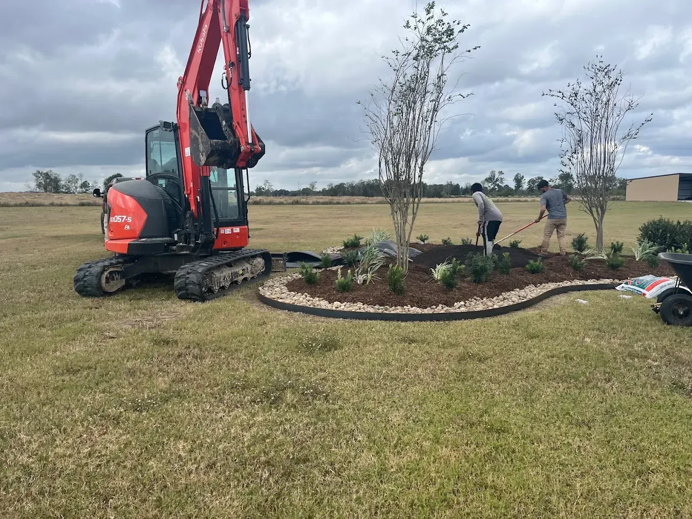 Small excavator beside a newly landscaped garden bed in a grassy field, with two workers planting trees under cloudy skies