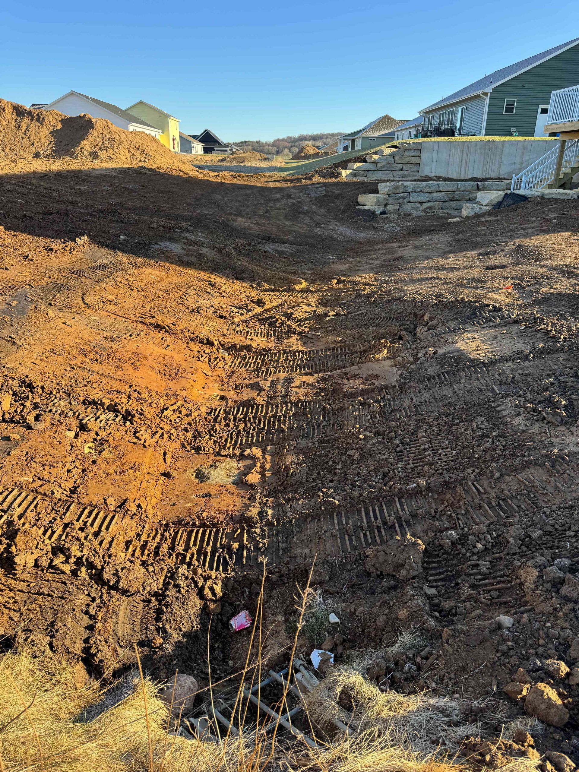 A construction site with a large pile of dirt, tire tracks, and a partially buried metal grate in the foreground.